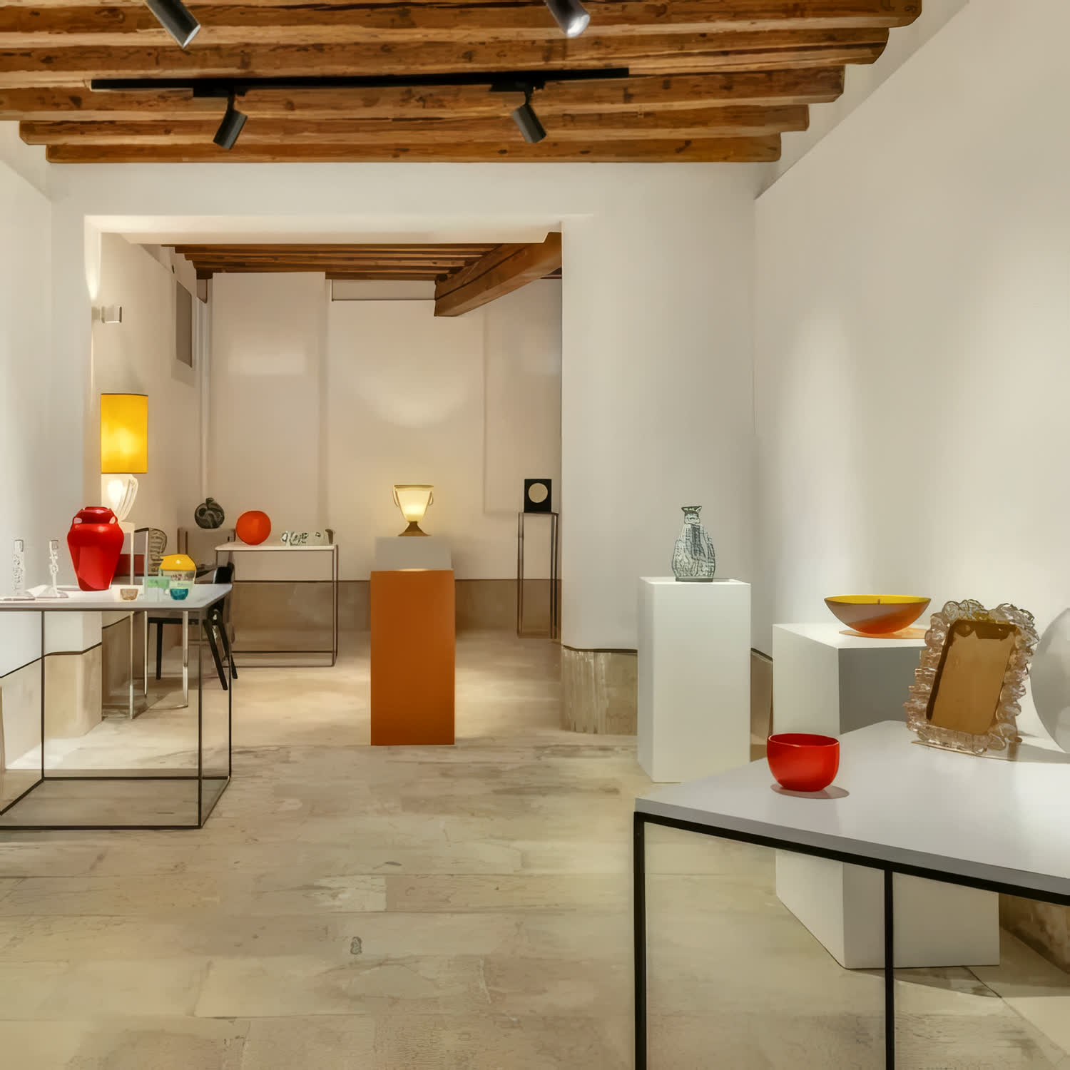 Sala della Galleria Diciotto a Venezia con le opere in vetro esposte su piedistalli bianchi, sotto travi di legno
